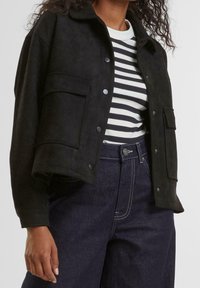 Veste en daim noire avec une fermeture à boutons, un col et une poche poitrine, associée à une chemise rayée noire et blanche et un pantalon en denim foncé.