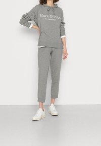 Felpa grigia in cotone con cappuccio e logo, abbinata a pantaloni della tuta svasati dello stesso colore e sneakers bianche su uno sfondo neutro.