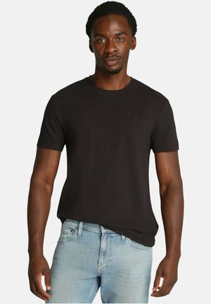 T-shirt nera a maniche corte in cotone, con scollo rotondo e un sottile dettaglio logo sul petto, abbinata a jeans azzurri chiari.