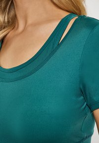 Teal színű, testhezálló felső sima, rugalmas anyagból. Kerek nyakú, és divatos vállkivágásokkal rendelkezik, megerősített varrásokkal.