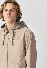 Jeune homme aux cheveux bouclés portant une veste à capuche beige clair avec fermeture éclair frontale et poches à rabat, fond neutre.