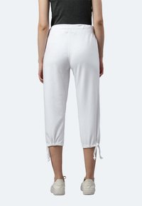 Weiße, knöchellange Jogginghosen mit einem elastischen Bund und verstellbaren Bändern an den Knöcheln. Weicher Stoff mit glattem Textur, kombiniert mit weißen Sneakers.