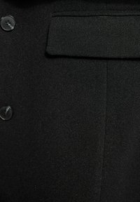 Primo piano di un tessuto di cappotto di lana nera con due bottoni neri e una tasca a pattina sul davanti.