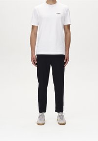 Vit bomullströja med rund halsringning och subtil logotyp. Kombinerad med svarta byxor och ljusgrå sneakers, stående på en enkel vit bakgrund.