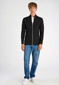 Svart zip-up tröja med hög krage, strukturerad stickdesign, slim fit och ribbade ärmar, ihop med ljusblå jeans och vita sneakers.