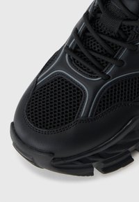 Black athletic shoe featuring a textured upper with mesh panels, grey accents, and a thick, patterned sole for support and traction.