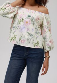 Blusa floreale con design off-shoulder, caratterizzata da fiori rosa e viola pastello su uno sfondo crema, maniche a sbuffo e texture simile al lino.