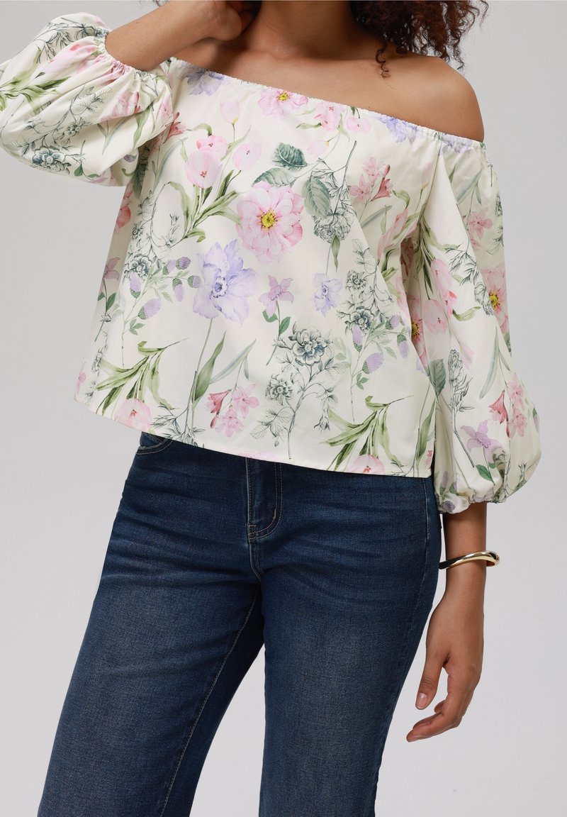 Blusa floreale con design off-shoulder, caratterizzata da fiori rosa e viola pastello su uno sfondo crema, maniche a sbuffo e texture simile al lino.