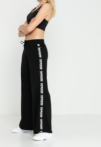 Pantalones de pierna ancha negros, confeccionados en una tela suave, que presentan el texto blanco "Reebok" a lo largo del costado. Incluyen una cintura con cordón ajustable.