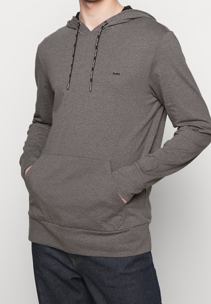 Homme portant un sweat à capuche gris avec des cordons noirs et une poche avant, assorti à un jean bleu foncé. Mains dans les poches.