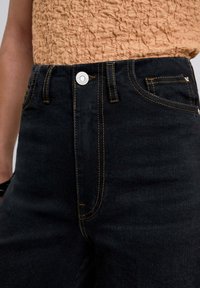 Hochtaillierte schwarze Jeans aus Denim mit kontrastierender gelber Stecknaht, silbernen Knopfverzierungen und klassischem Fünf-Taschen-Design.