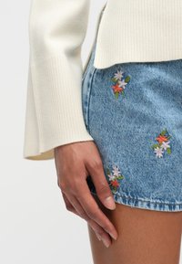 Kremfarget ribbestrikket langermet topp kombinert med blå denimshorts med flerfarget blomsterbroderi og frynsete kant, som viser en avslappet stil.