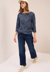 Frau mit einer weißen Mütze, einem blauen gemusterten 3/4-Ärmel-Oberteil, dunkelblauen Weit-Leg-Jeans und cremefarbenen Turnschuhen, die vor einem beigen Hintergrund steht.