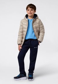 Bēša pūderveida jaka ar kapuci, zils džemperis, tumši zili sporta bikses un tumši zili sporta apavi ar ziliem akcentiem. Vienkāršs logotips uz apģērba.