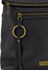Schwarzer Leder-Rucksack mit strukturiertem Finish, zwei Reißverschlüssen in Goldoptik, einem runden goldfarbenen Hardware-Akzent und einem Namensschild.