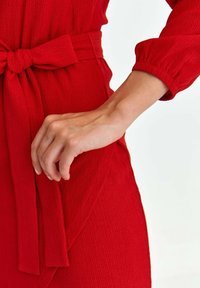 Robe rouge texturée avec un détail de taille nouée et de longues manches légèrement bouffantes. Le tissu semble doux avec un motif subtil.