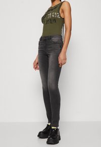 Grönribbad tanktop med grafisk tryck, kombinerad med mörkgrå skinnyjeans och kraftiga svarta skor, som visar en modern avslappnad stil.