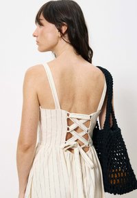 Trägerloses cremefarbenes Kleid mit vertikalen Streifen, Schnürdesign auf der Rückseite und einem ausgestellt geschnittenen Rock. Eine schwarze geflochtene Handtasche sorgt für einen Kontrast.