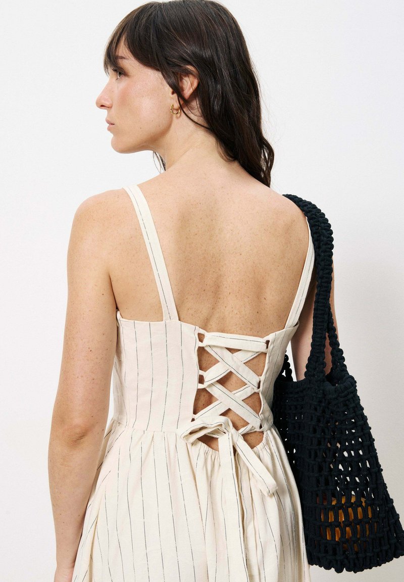 Trägerloses cremefarbenes Kleid mit vertikalen Streifen, Schnürdesign auf der Rückseite und einem ausgestellt geschnittenen Rock. Eine schwarze geflochtene Handtasche sorgt für einen Kontrast.
