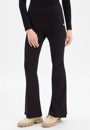 Donna che indossa pantaloni neri a vita alta svasati con cuciture verticali, abbinati a mocassini beige con suola robusta e top nero aderente.