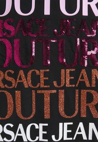 Sort stof med lagdelt tekst i forskellige skrifttyper og farver: pink pailletter, glitrende bronze og hvid. Teksten indeholder "VERSACE JEANS COUTURE."