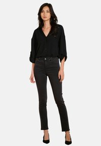 Mujer de pie con una camisa negra holgada de botones, unos jeans ajustados negros de cintura alta con detalles de tachuelas, y tacones negros de punta.