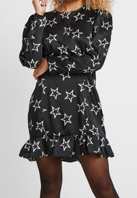 Svart klänning med långa ärmar, vitt stjärnmönster, rund halsringning och volangkant. Tillverkad av ett slätt tyg med en lätt glans.