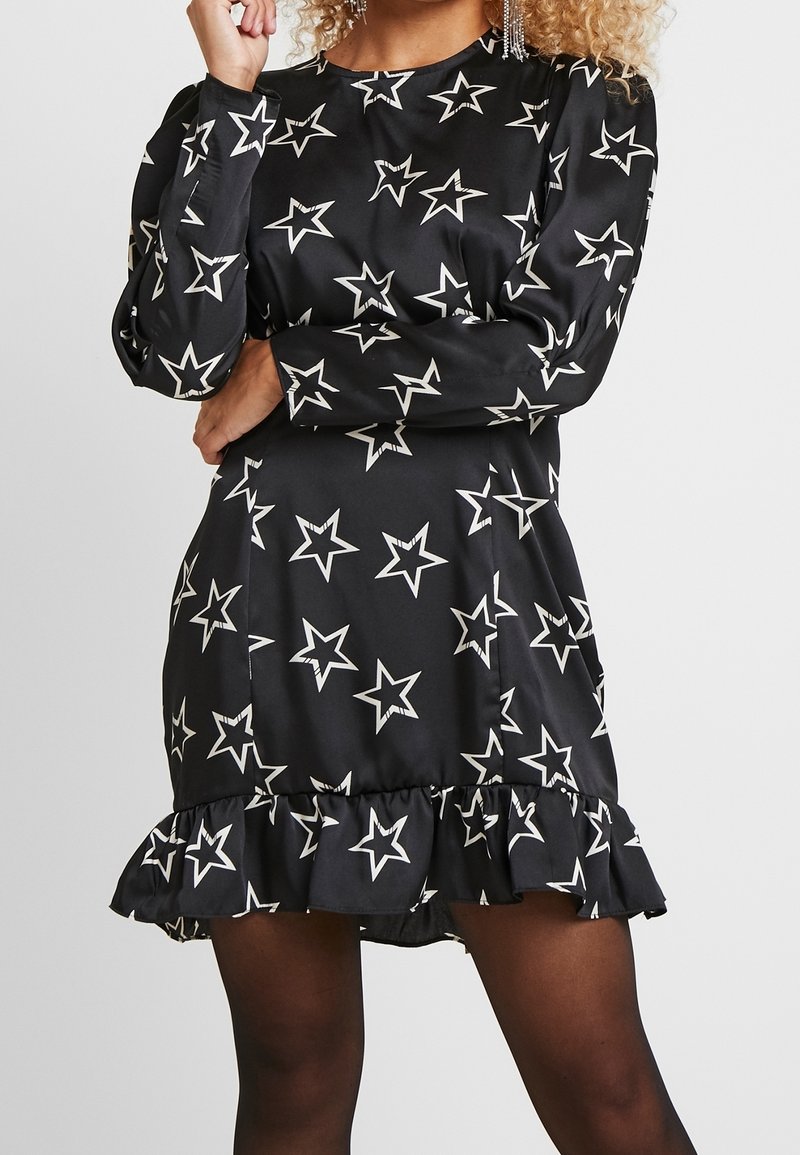 Svart klänning med långa ärmar, vitt stjärnmönster, rund halsringning och volangkant. Tillverkad av ett slätt tyg med en lätt glans.