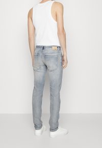 Pantalones vaqueros de denim azul claro con un corte ajustado, que presentan un parche de cuero en la parte trasera de la cintura y dos bolsillos traseros, combinados con una camiseta sin mangas blanca.