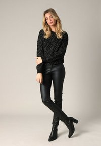 Femme aux cheveux blonds portant un pull noir à motifs, un pantalon en cuir noir et des bottines à talons noires, se tenant les jambes croisées.