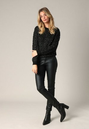 Femme aux cheveux blonds portant un pull noir à motifs, un pantalon en cuir noir et des bottines à talons noires, se tenant les jambes croisées.