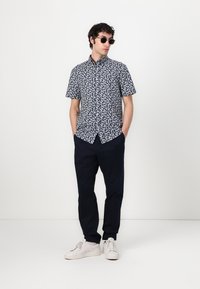 Man met zonnebril, kortemouwen marineblauw shirt met wit bladerenpatroon, marineblauwe broek en witte sneakers, staand met handen in de zakken.