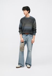 Siv in modro gradientno pleten pulover, svetlo modre kavbojke z širokimi hlačnicami, rjav pas, drži sivo kapo, bele in rjave superge. Preprosto ozadje.