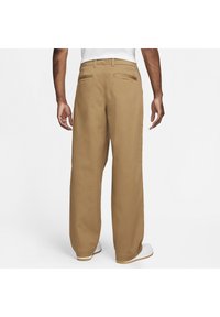 Man med löst sittande beige byxor och vita sneakers, stående med ryggen mot kameran mot en enkel vit bakgrund.