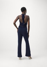 Mörkblå jumpsuit med halterneck och öppen rygg, som har en åtsittande bodice och utsvängda byxor. Slät textur, minimalistisk design.