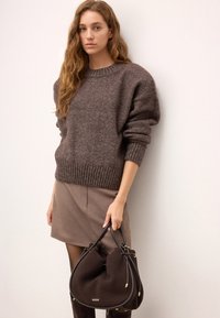 Brun, stickad tröja med rund halsringning och ribbade ärmar, kombinerad med en beige kjol. En mörkbrun väska med guldhardware hålls av modellen.