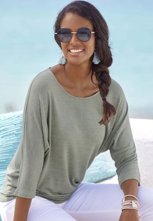 Helles grünes, locker sitzendes Oberteil mit 3/4-Ärmeln, kombiniert mit weißen Hosen. Die Accessoires umfassen runde Sonnenbrillen und hellblaue Quastenohrringe.
