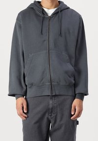 Uomo che indossa una felpa con zip grigia scura con tasche frontali e pantaloni grigi scuri abbinati, in piedi contro uno sfondo chiaro e uniforme.