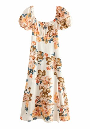 Femme portant une robe blanche à fleurs avec des manches bouffantes et des chaussures à bride à la cheville, debout près d'une colonne blanche en plein air à la lumière du jour.