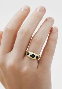 Anillo de oro con tres piedras negras, con un acabado suave y pulido y una banda redondeada. Lleva puesto en una mano de tono de piel claro.