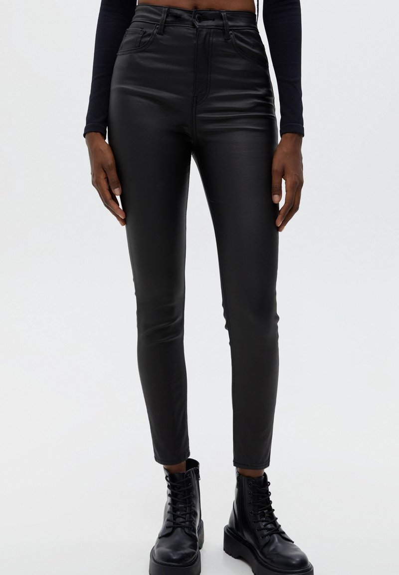 Personne portant un pantalon skinny taille haute en simili cuir noir et des bottines noires à lacets, debout devant un fond clair uni.