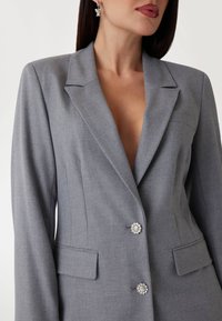 Blazer grigio su misura con scollo a V profondo, due tasche frontali e bottoni metallici floreali. La texture del tessuto appare liscia e strutturata.