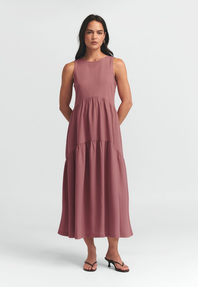 Maksi obleka iz svetlo mauve tkanine s brezrokavnim dizajnom, okroglim ovratnikom in plastenim krilom. Nosi se z črnimi sandali na peto.