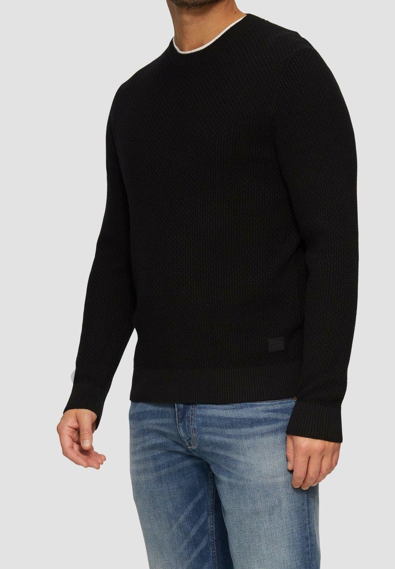 Homme portant un pull noir en maille texturée avec des poignets et un ourlet côtelés, associé à un jean denim bleu clair sur un fond gris uni.