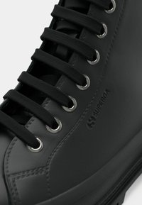 Prim-plan al unei pantof de piele negre cu șireturi negre trecute prin ochiurile metalice argintii și logo-ul "Superga" embossat pe lateral.