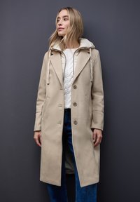 Beiger Mantel mit Kapuze und Knöpfen vorne, aus weichem Material mit großen Taschen, getragen über einem weißen strukturierten Pullover und blauen Jeans.