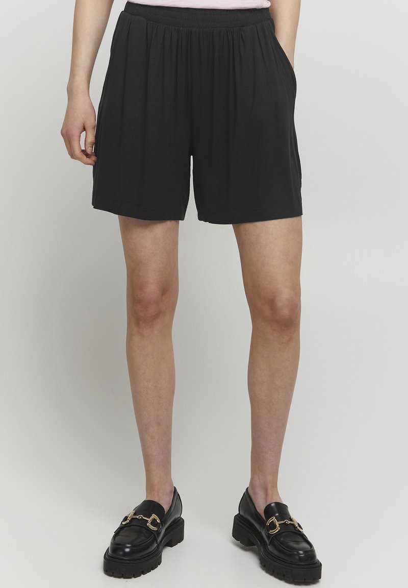 Svarta shorts med elastisk midja, löst sittande, slät textur och sidofickor. Kombination med glänsande svarta loafers med guldaccenter.