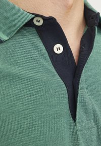Grøn polo skjorte med tekstureret stof, marineblå krave og to lyse knapper. Kraven har en kontrastfarvet ribbet tekstur.
