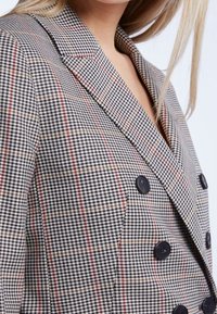 Blazer croisé à motif pied-de-poule avec des accents gris, rouge et crème. Il présente des boutons noirs et un col cranté.