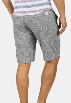 Homme portant un short gris texturé avec deux poches arrière boutonnées et une chemise blanche rayée rouge et bleue rentrée à l'intérieur.